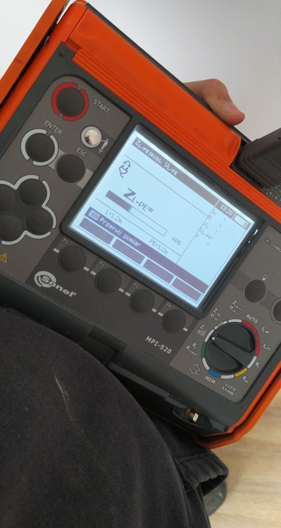Miernik elektryczny Sonel MPI-520 w trakcie pomiaru impedancji pętli zwarcia ZL-PE, wyświetlający wartości L=1,2m i PE=1,2m na ekranie LCD, trzymany w dłoni nad ciemnymi spodniami.
