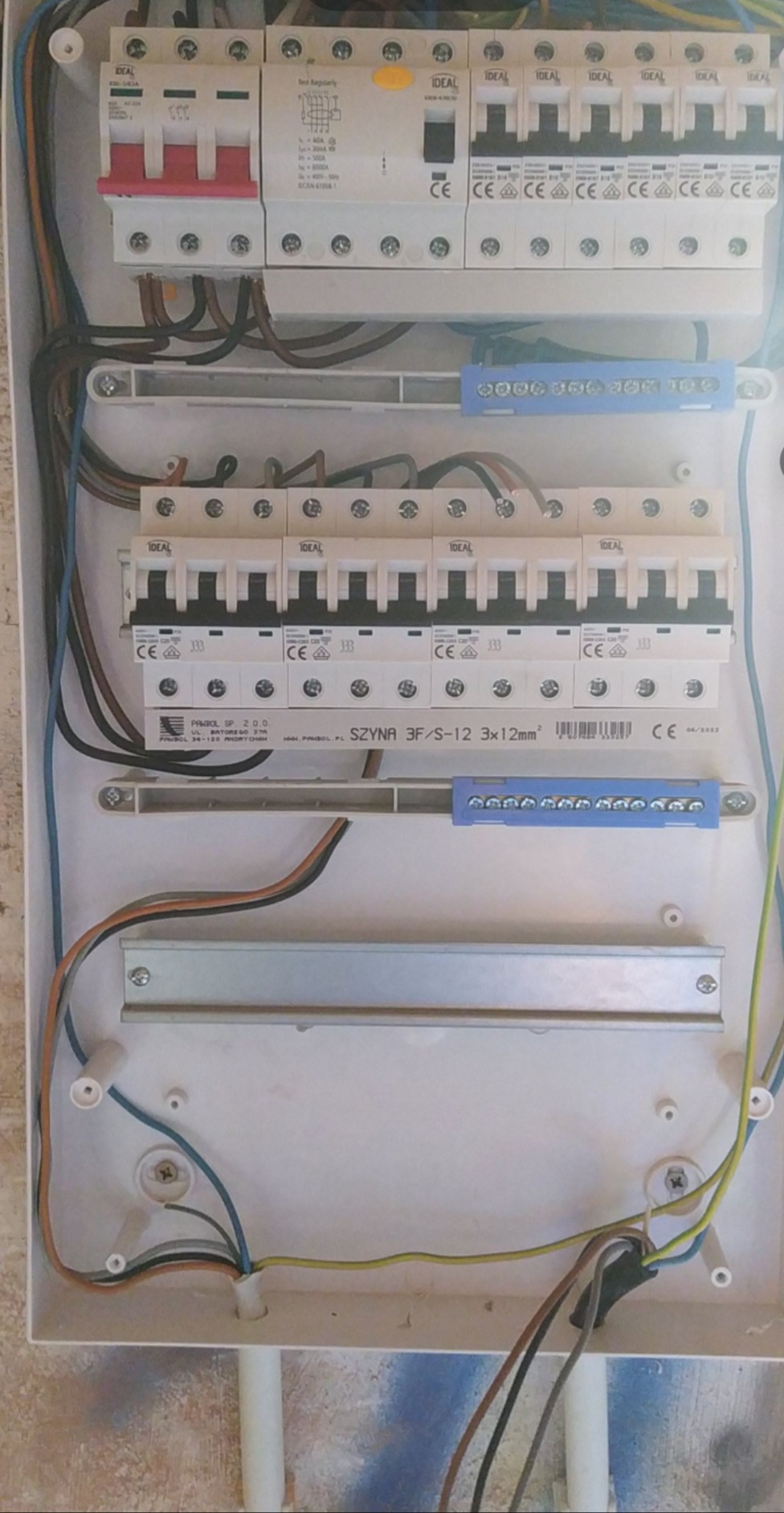 Otwarta skrzynka bezpiecznikowa z widocznymi bezpiecznikami, szynami DIN i przewodami elektrycznymi wewnątrz białej obudowy. Widoczne ślady po wierceniu w obudowie.