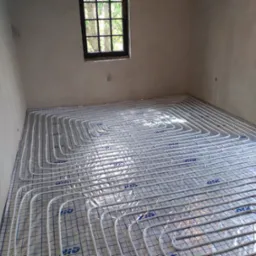 Białe rury ogrzewania podłogowego ułożone na folii z nadrukowaną siatką, widok z góry, surowe ściany w tle, okno z czarną ramą.