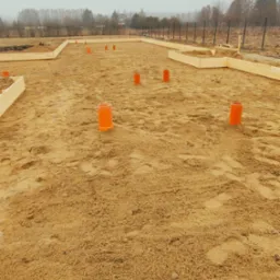Przygotowany teren pod fundamenty domu jednorodzinnego z wystającymi pomarańczowymi rurami instalacji w piasku, obramowany drewnianą szalówką, w tle ogrodzenie i drzewa w pochmurny dzień.