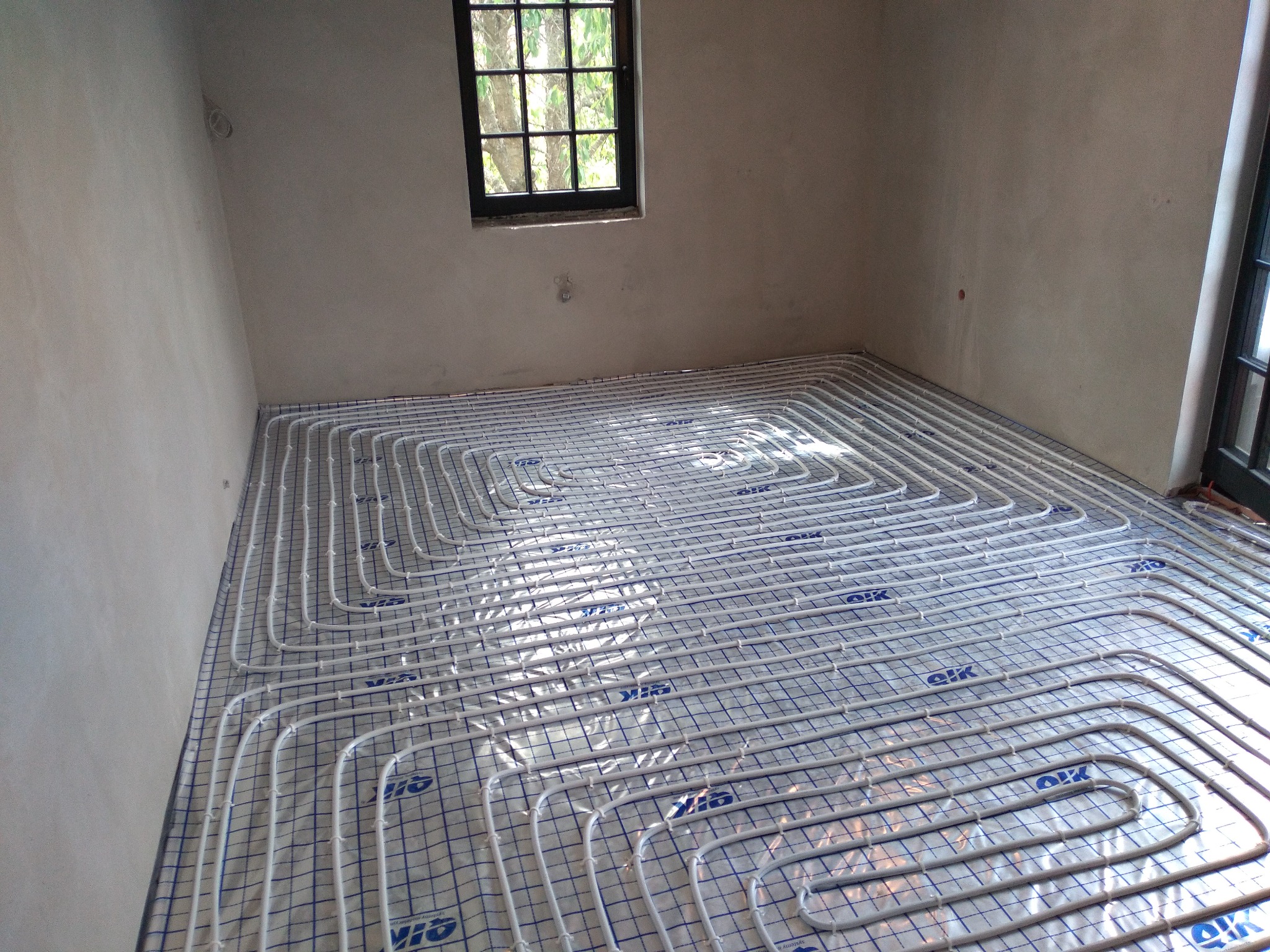 Białe rury ogrzewania podłogowego ułożone na folii z nadrukowaną siatką, widok z góry, surowe ściany w tle, okno z czarną ramą.