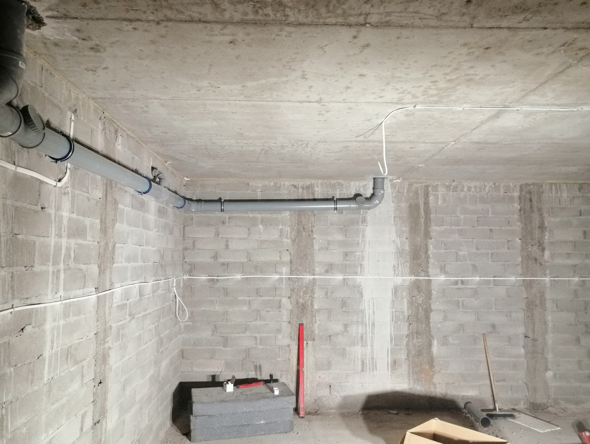 Szare rury kanalizacyjne podwieszone pod surowym betonowym sufitem w pomieszczeniu z bloczków betonowych, widoczne narzędzia i materiały budowlane na podłodze.