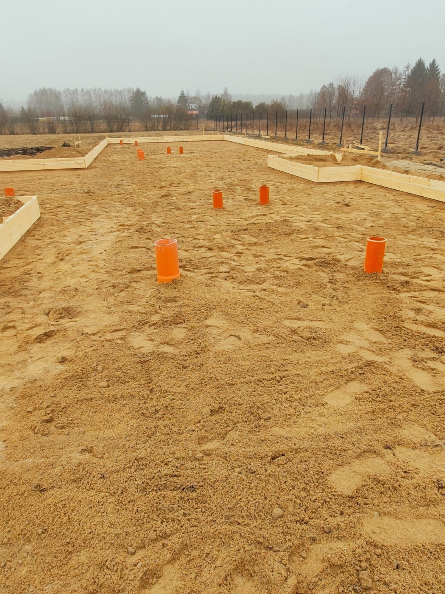 Przygotowany teren pod fundamenty domu jednorodzinnego z wystającymi pomarańczowymi rurami instalacji w piasku, obramowany drewnianą szalówką, w tle ogrodzenie i drzewa w pochmurny dzień.