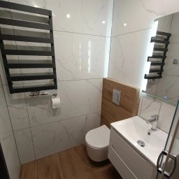 Dream House Antoni Ostrowski - Nowoczesna łazienka z białymi marmurkowymi płytkami, drewnianą obudową toalety, czarnym grzejnikiem drabinkowym i białą umywalką z szafką.