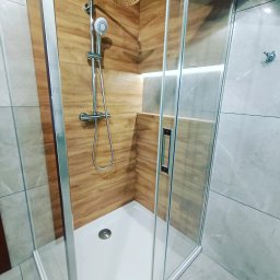 Dream House Antoni Ostrowski - Kabina prysznicowa z drewnopodobnymi płytkami ściennymi i szarymi płytkami na ścianach, z oświetleniem LED w niszy.