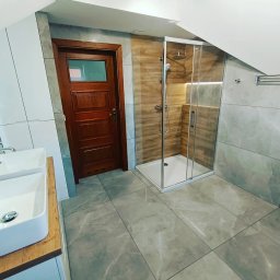 Dream House Antoni Ostrowski - Nowoczesna łazienka we Wrocławiu z kabiną prysznicową typu walk-in, dwiema umywalkami nablatowymi na drewnianym blacie i ciemnymi drewnianymi drzwiami, wykończona płytkami w odcieniach szarości...