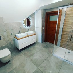 Dream House Antoni Ostrowski - Nowoczesna łazienka z szarymi płytkami, podwójną białą umywalką na drewnianym blacie, okrągłym lustrem z podświetleniem LED oraz kabiną prysznicową z drewnopodobnymi płytkami wewnątrz