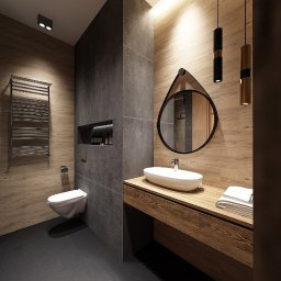Dream House Antoni Ostrowski - Nowoczesna łazienka z drewnopodobnymi płytkami na ścianach, szarym wykończeniem, okrągłym lustrem zawieszonym na skórzanym pasku, wiszącą toaletą i grzejnikiem drabinkowym.
