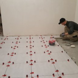 Dream House Antoni Ostrowski - Układanie płytek podłogowych z użyciem systemu poziomowania, widoczny pracownik w rękawicach i zacierka, przygotowane płytki i wiaderko z klipsami.