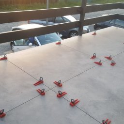 Dream House Antoni Ostrowski - Szare płytki na balkonie w trakcie układania, użyte czerwone klipsy poziomujące, w tle zaparkowane samochody i balustrada