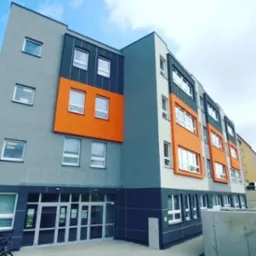 Wielopiętrowy budynek we Wrocławiu z szarą elewacją, pomarańczowymi akcentami i białymi ramami okiennymi, widziany z perspektywy lekko od dołu.