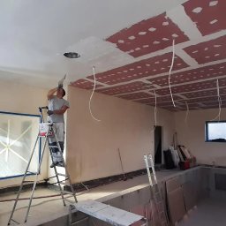 Dream House Antoni Ostrowski - Pracownik na drabinie szpachluje sufit z płyt karton gips, widoczne przewody oświetleniowe i częściowo pomalowane fragmenty sufitu.