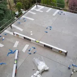 Balkon w trakcie układania płytek: poziomice, krzyżyki dystansowe i taśma maskująca na szarym podłożu, widok z góry na fragment balkonu z metalową barierką i zielenią w tle.