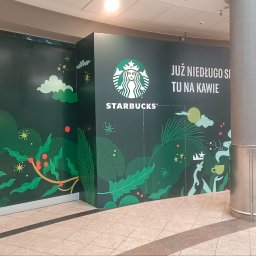 Dream House Antoni Ostrowski - Oklejona ścianka działowa z grafiką Starbucks w galerii handlowej, z napisem 'Już niedługo się tu napijesz kawy'. W tle przechodzący ludzie.