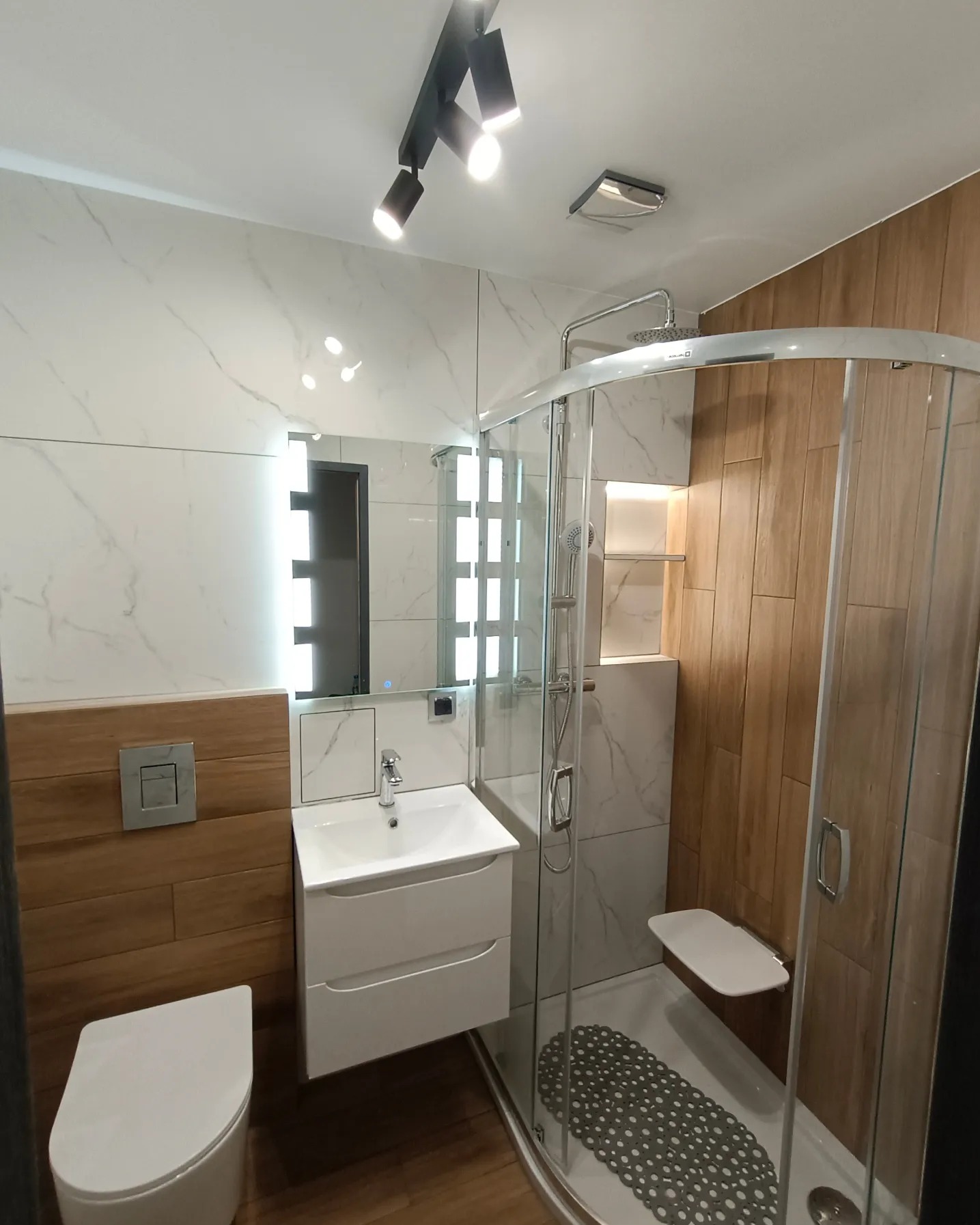 Nowoczesna łazienka z kabiną prysznicową z siedziskiem, umywalką z szafką i podświetlanym lustrem, wykończona płytkami imitującymi marmur i drewno