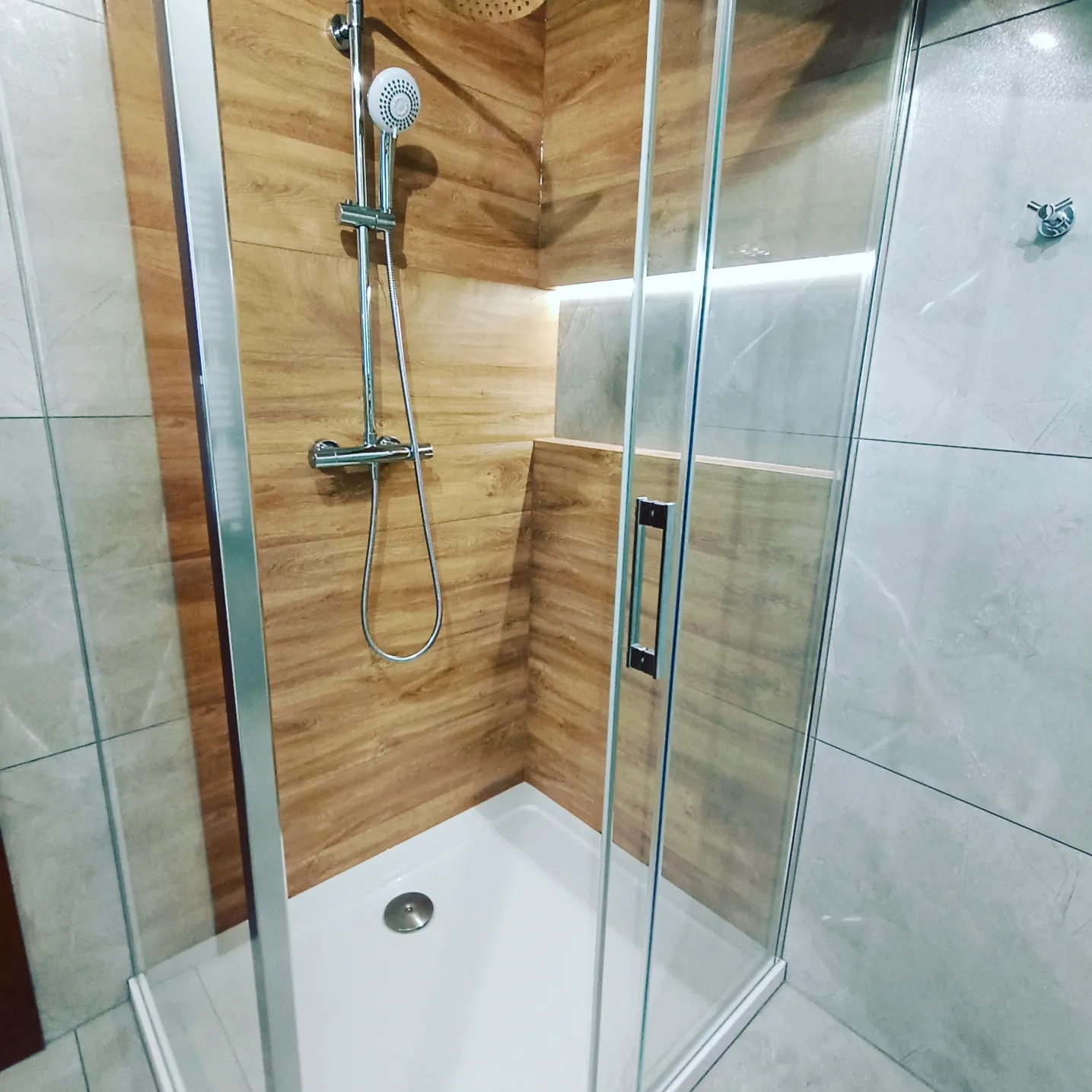 Kabina prysznicowa z drewnopodobnymi płytkami ściennymi i szarymi płytkami na ścianach, z oświetleniem LED w niszy.