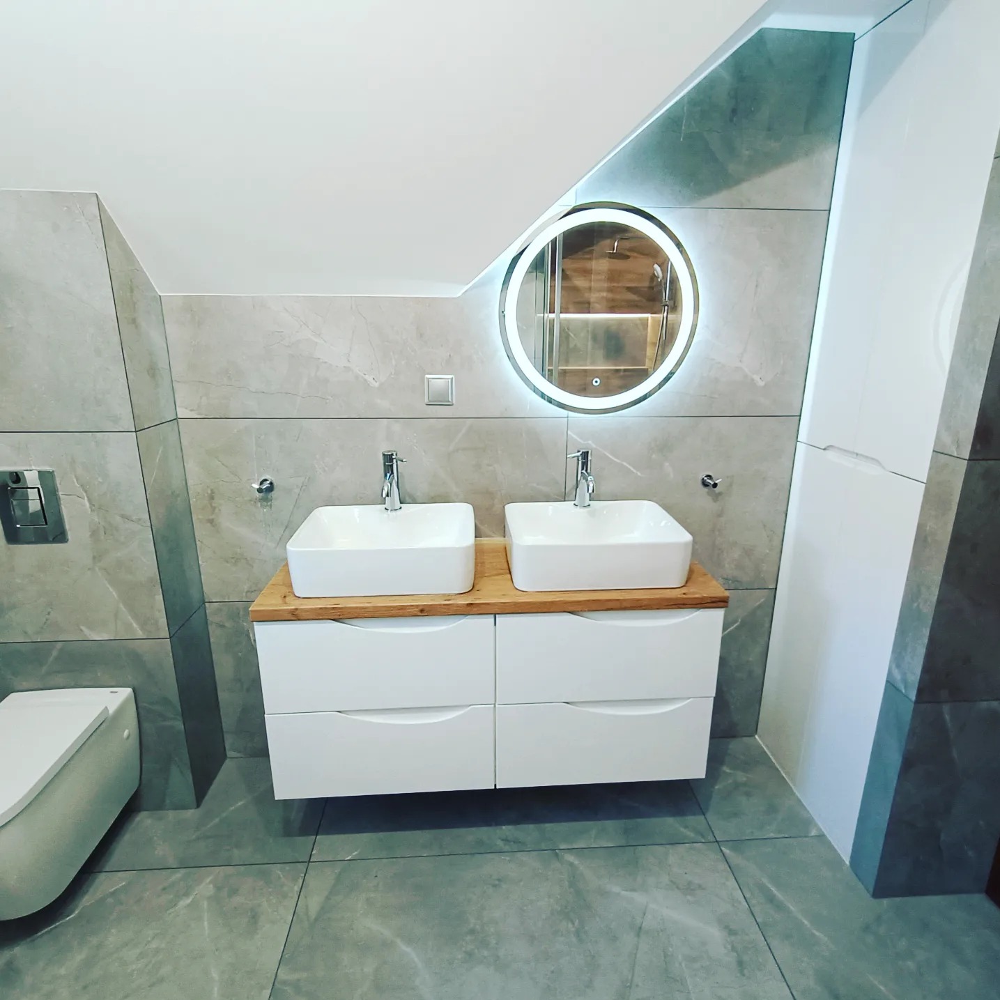 Nowoczesna łazienka na poddaszu z dwoma białymi umywalkami nablatowymi na drewnianym blacie, białymi szafkami pod umywalkami, okrągłym lustrem z podświetleniem LED i szarymi płytkami imitującymi...