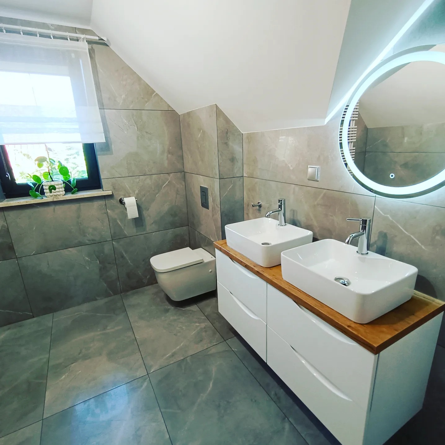 Nowoczesna łazienka na poddaszu z dwoma kwadratowymi umywalkami nablatowymi na białej szafce z drewnianym blatem, podświetlane okrągłe lustro, szare płytki imitujące marmur na ścianach i podłodze...