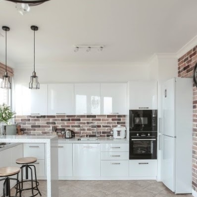 Nowoczesna, jasna kuchnia z białymi, lakierowanymi szafkami, ceglaną ścianą nad blatem, wyspą kuchenną z dwoma hokerami i wiszącymi lampami w stylu industrialnym.