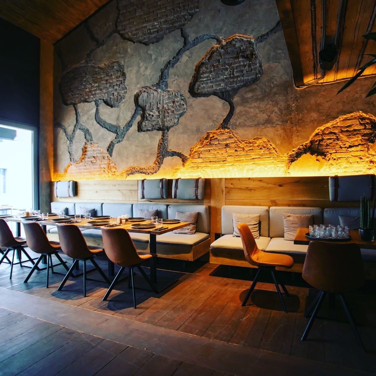 Wnętrze restauracji z dekoracyjną ścianą imitującą odsłoniętą cegłę i podświetleniem, drewnianą boazerią, szarą tapicerowaną ławą i brązowymi krzesłami przy drewnianych stołach z nakryciami.