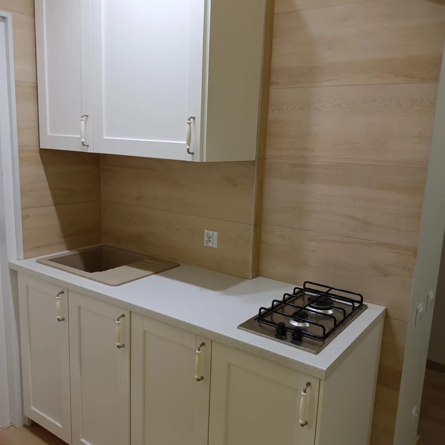 Nowoczesna kuchnia z białymi szafkami z kremowymi uchwytami, drewnopodobną okładziną ścienną, beżowym zlewem i dwupalnikową płytą gazową.