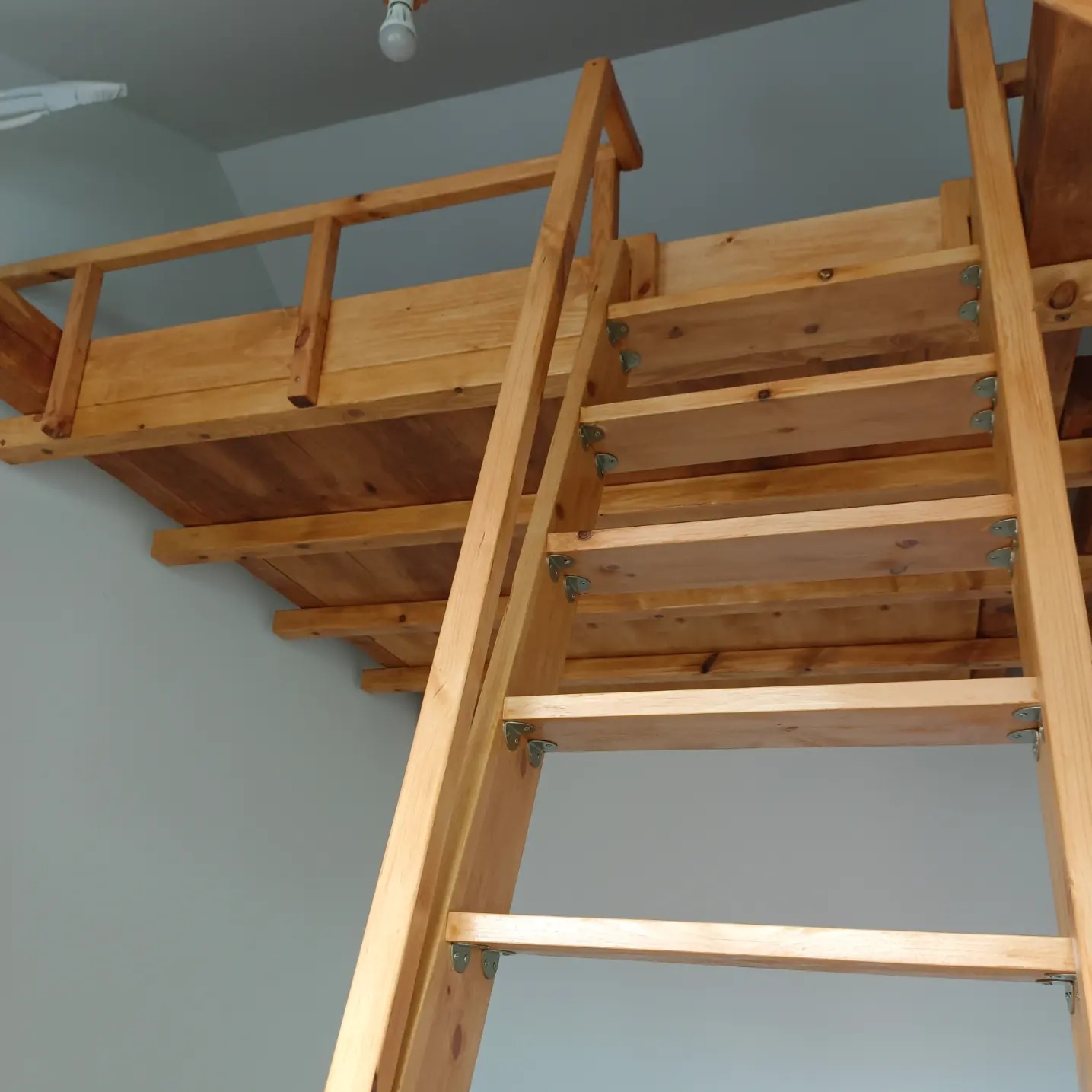 Drewniana konstrukcja antresoli z drabinką, widok z dołu ukazujący surowy stan drewna i metalowe mocowania stopni.