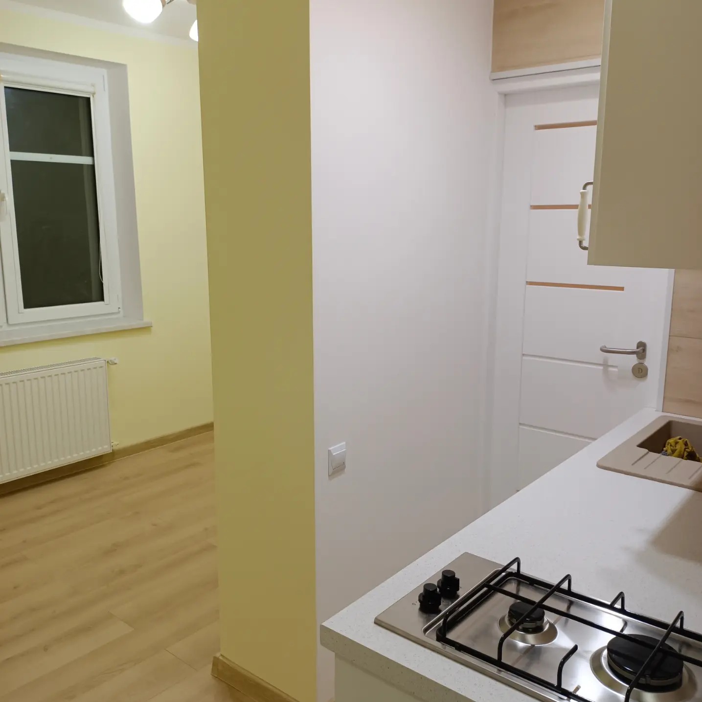 Fragment wnętrza: widok na kuchnię z płytą gazową, zlewozmywakiem, białą ścianą i drzwiami wewnętrznymi oraz fragmentem pokoju z panelami podłogowymi i oknem.