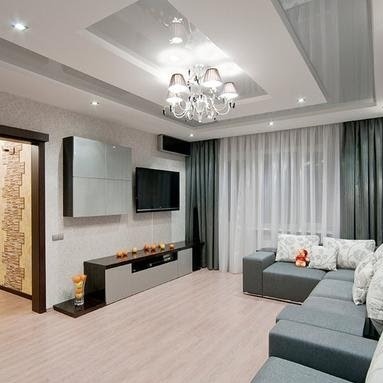 Nowoczesny salon z szarym narożnikiem, białymi zasłonami i podwieszanym sufitem z oświetleniem punktowym. Na ścianie szafki w kolorze szarym i czarnym oraz telewizor. Podłoga wyłożona panelami...