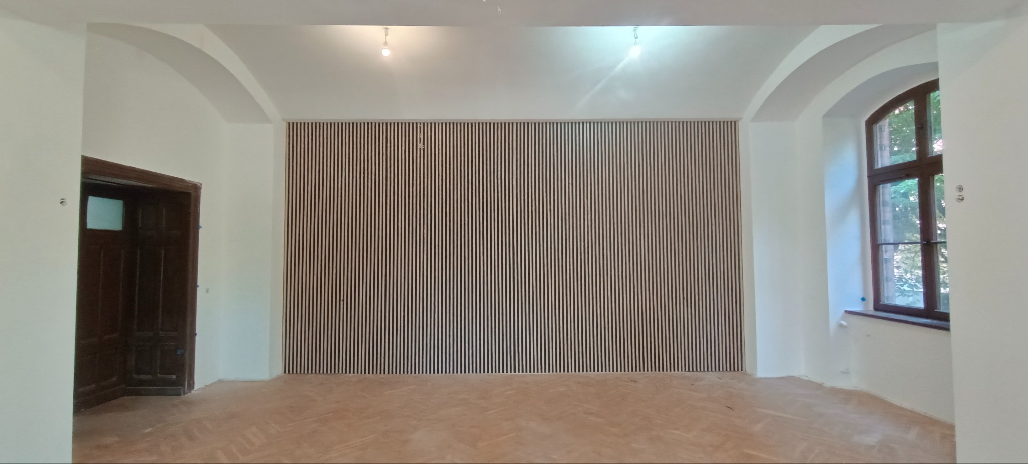 Wnętrze po montażu drewnianych lameli na ścianie, widoczne drzwi ukryte i fragment parkietu. Białe ściany z łukowymi przejściami i oknem.