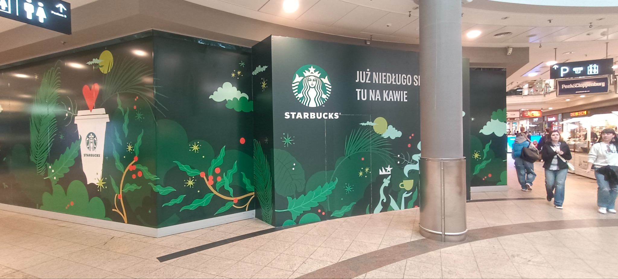 Oklejona ścianka działowa z grafiką Starbucks w galerii handlowej, z napisem 'Już niedługo się tu napijesz kawy'. W tle przechodzący ludzie.