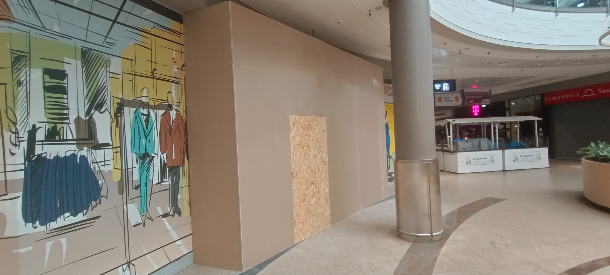 Zamurowane wejście do lokalu w galerii handlowej, z widocznym fragmentem dekoracji ściennej przedstawiającej odzież. Częściowo zasłonięte przez filar.