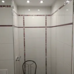 Wnętrze wykończonej łazienki z białymi płytkami, ozdobnymi bordiurami w odcieniach fioletu i brązu. Ustawione krzesło z brązowym siedziskiem.