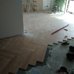 MARIO-REM Mariusz Skonieczny - Układanie parkietu w jodełkę. Widoczne narzędzia, poziomica, płytki z dystansami. W tle drabiny i resztki materiałów budowlanych, co wskazuje na trwający remont.