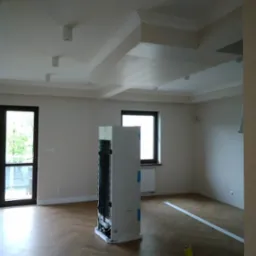 Jasne wnętrze po remoncie: parkiet w jodełkę, świeżo pomalowane ściany, ozdobny sufit z listwami. Widoczne okna i drzwi balkonowe. Na podłodze stoi niezapakowana lodówka.