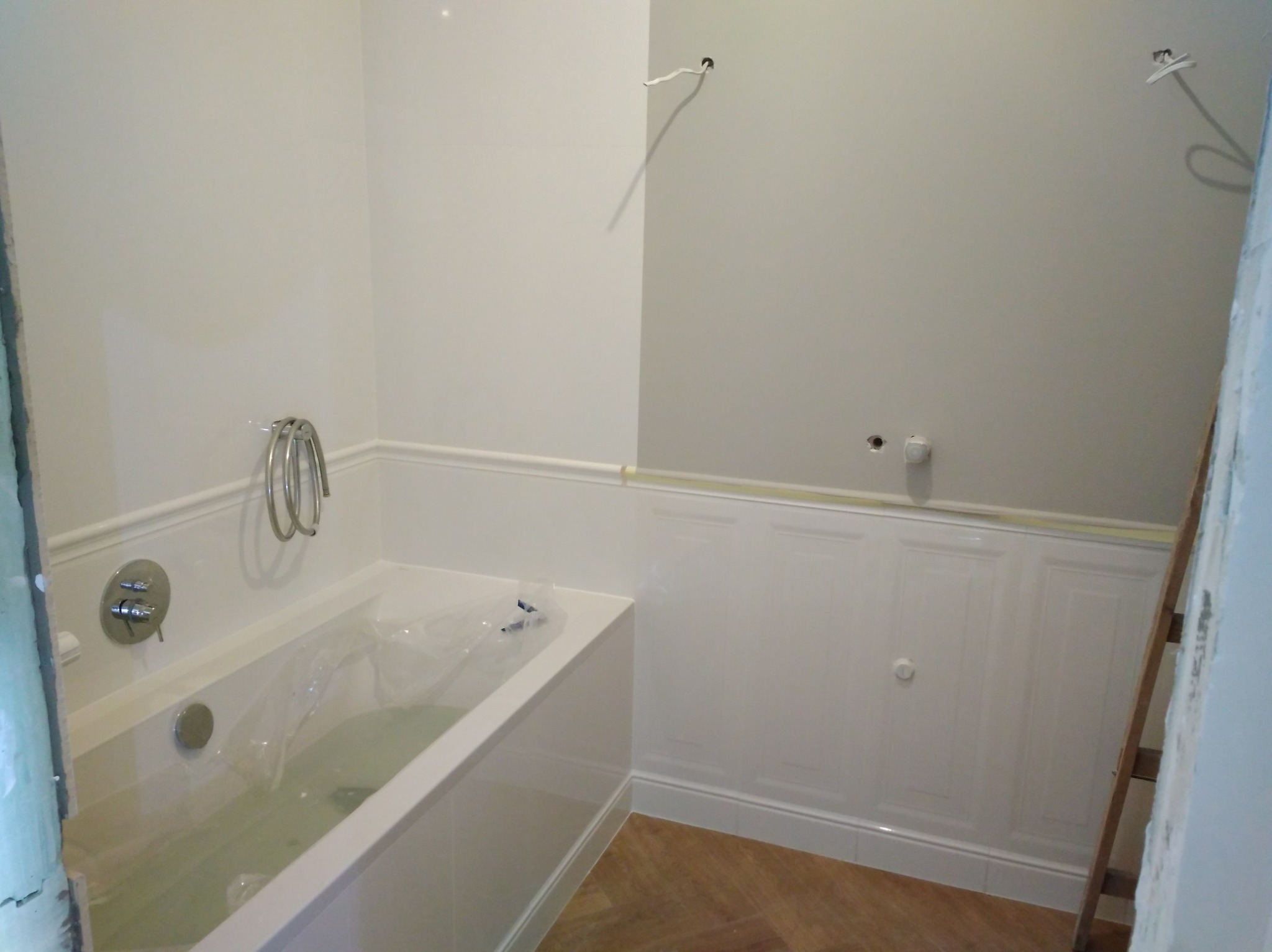 Biała wanna z wodą i plastikową folią zabezpieczającą, obok ściana z panelami dekoracyjnymi. Widoczna drabina i wystające kable.