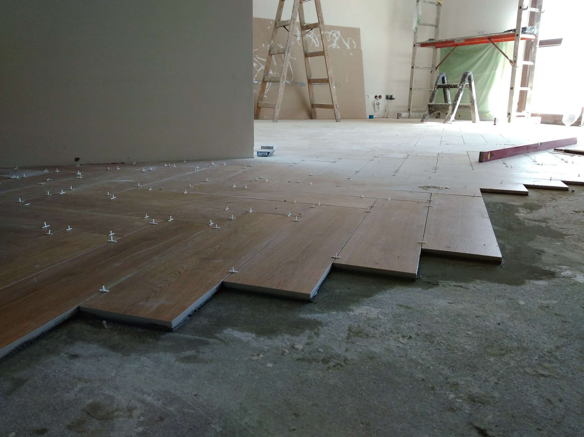 Układanie drewnopodobnych płytek na podłodze z użyciem krzyżyków dystansowych. W tle drabiny malarskie i przygotowane ściany do dalszych prac.