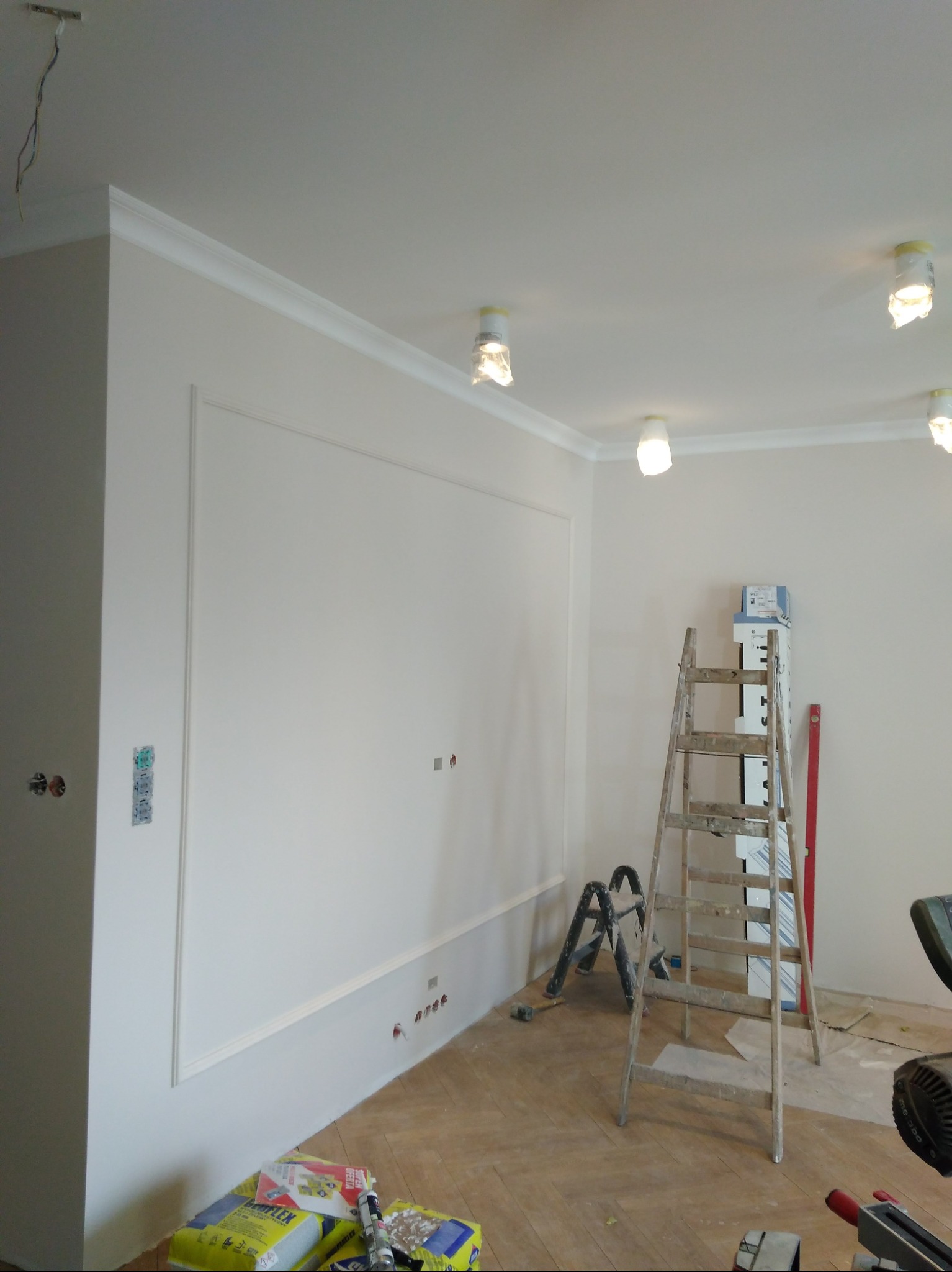 Białe ściany z dekoracyjnymi listwami i sztukaterią, drewniana podłoga, drabina i narzędzia. Remont wnętrza w trakcie realizacji, oświetlenie zabezpieczone folią.