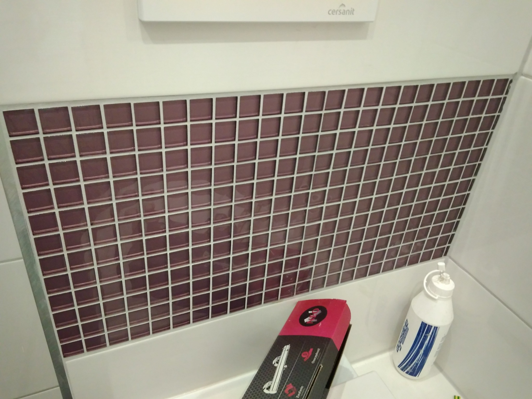 Bordowa mozaika w łazience, ułożona na ścianie nad wanną. Widoczne narzędzia glazurnicze i biała fuga. Cersanit.