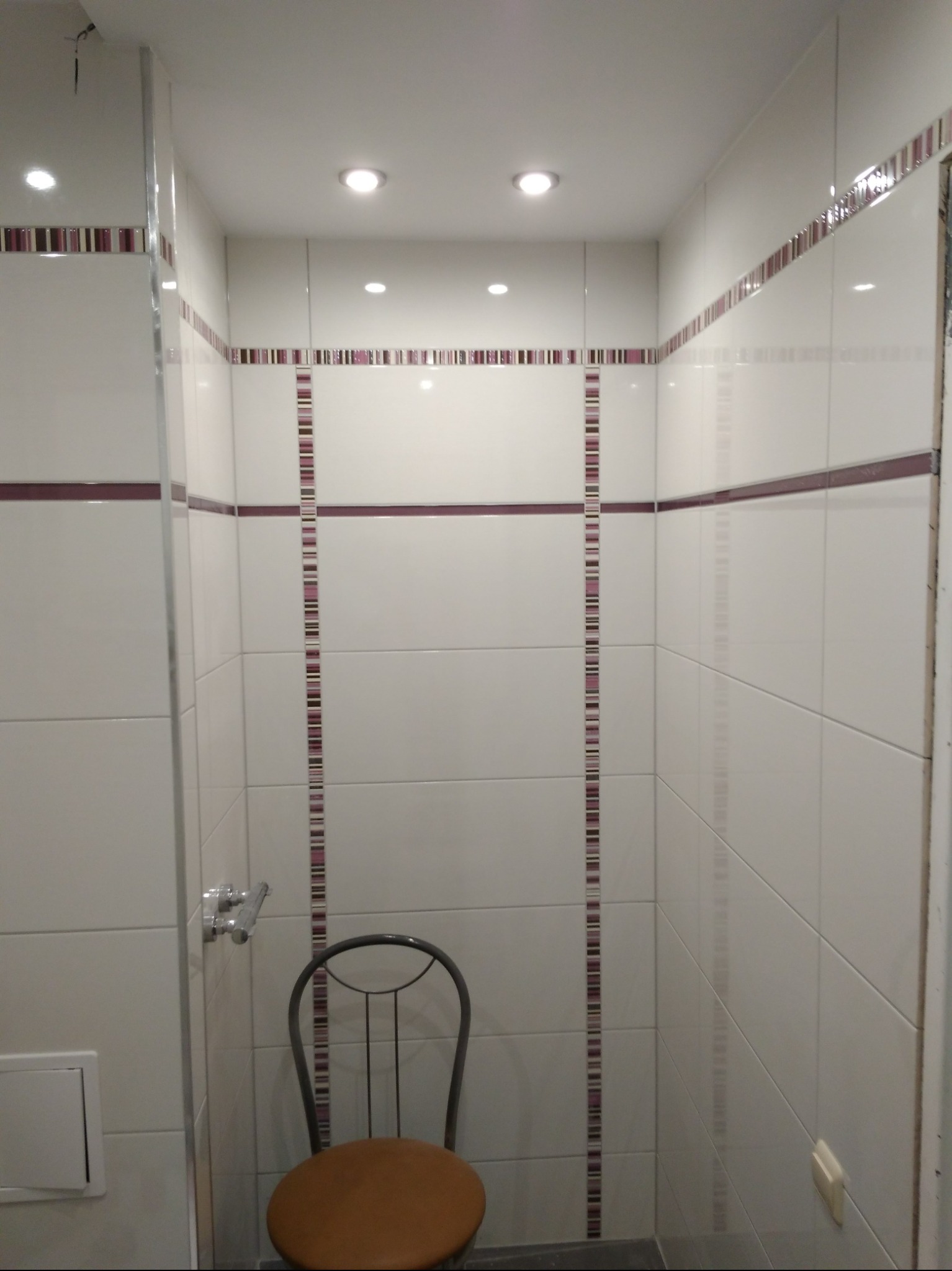Wnętrze wykończonej łazienki z białymi płytkami, ozdobnymi bordiurami w odcieniach fioletu i brązu. Ustawione krzesło z brązowym siedziskiem.