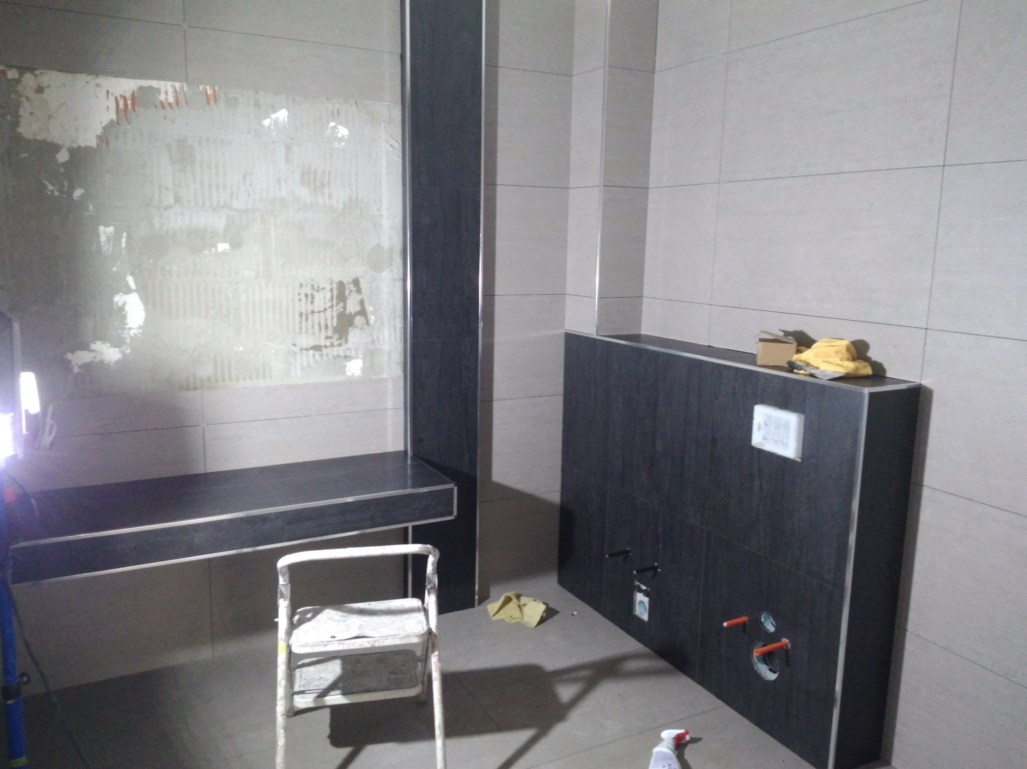 Łazienka w trakcie prac: częściowo położone płytki w odcieniach szarości, widoczny stelaż podtynkowy WC, drabina malarska i przygotowane miejsce pod umywalkę.