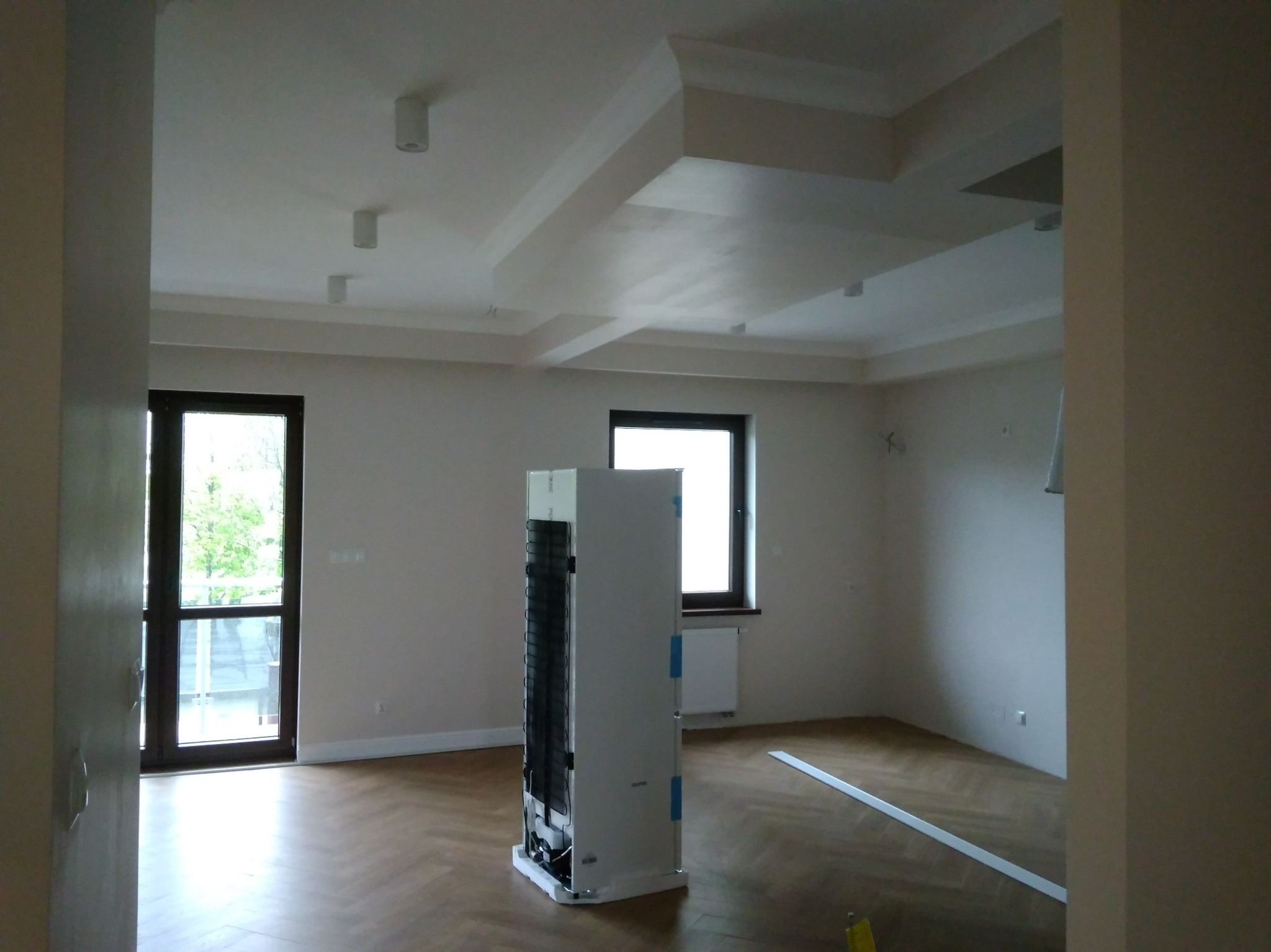 Jasne wnętrze po remoncie: parkiet w jodełkę, świeżo pomalowane ściany, ozdobny sufit z listwami. Widoczne okna i drzwi balkonowe. Na podłodze stoi niezapakowana lodówka.