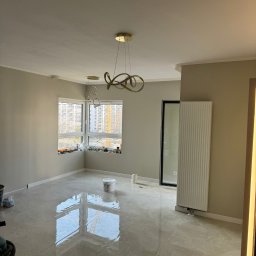 DONE PL SPÓŁKA Z OGRANICZONĄ ODPOWIEDZIALNOŚCIĄ - Pomalowane na szaro wnętrze pokoju z połyskującą podłogą z płytek, widoczne okno z widokiem na budowę, designerskie lampy sufitowe i biały grzejnik.