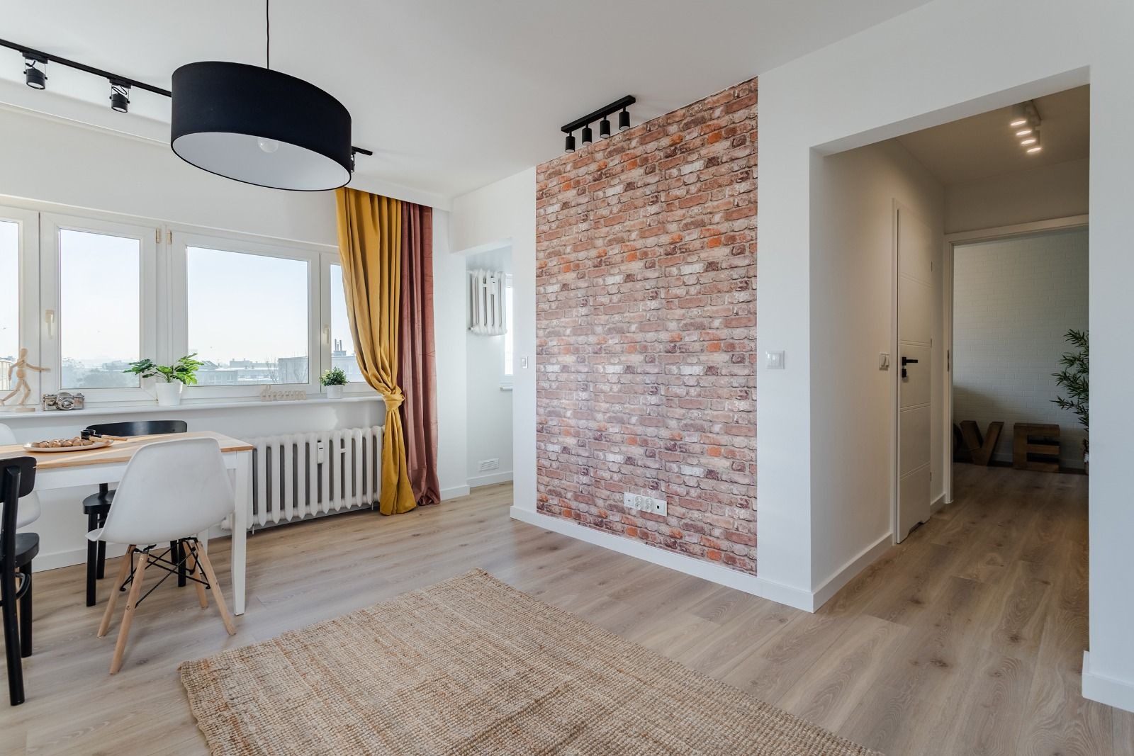 Jasne wnętrze mieszkania z drewnianą podłogą, ceglaną ścianą akcentującą, białym stołem z krzesłami i dużym oknem z zasłonami w odcieniach złota i brązu.