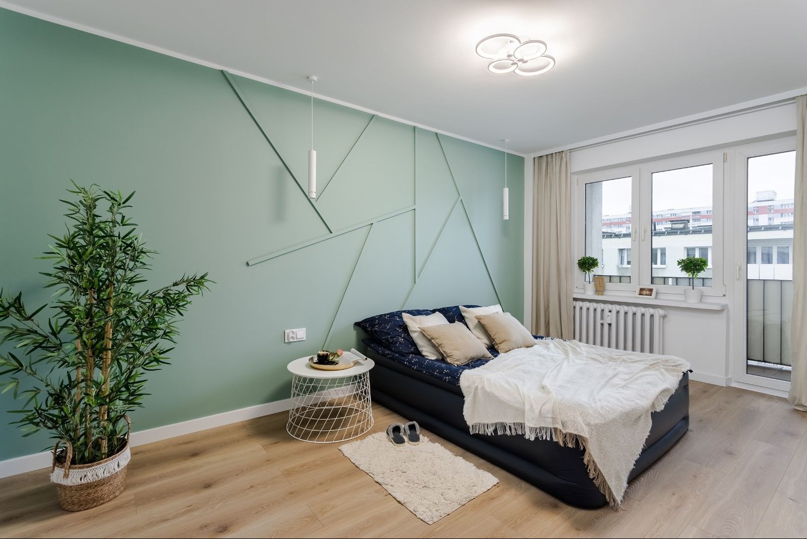 Jasna sypialnia z pistacjową ścianą zdobioną geometrycznymi listwami, łóżko z granatową pościelą i beżowymi poduszkami, biała lampa sufitowa i wiszące lampy cylindryczne, drewniana podłoga...