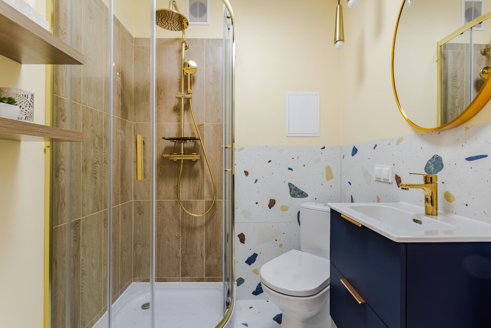 Jasna łazienka z kabiną prysznicową, złotą armaturą i umywalką w szafce w kolorze granatowym, ściany wykończone płytkami imitującymi lastryko.