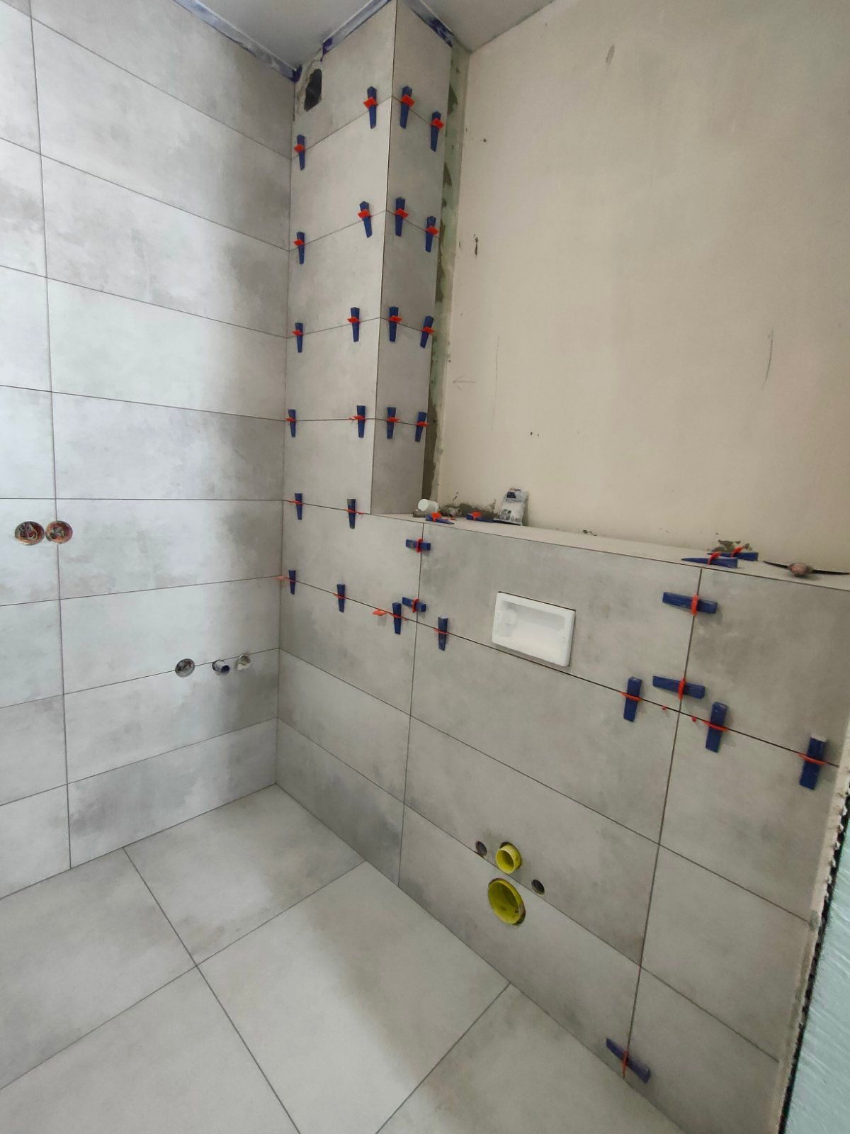 Świeżo ułożone, poziome, szare płytki w łazience, zabezpieczone niebieskimi klipsami poziomującymi i czarną żyłką; widoczne otwory na instalacje i fragment ściany bez płytek.