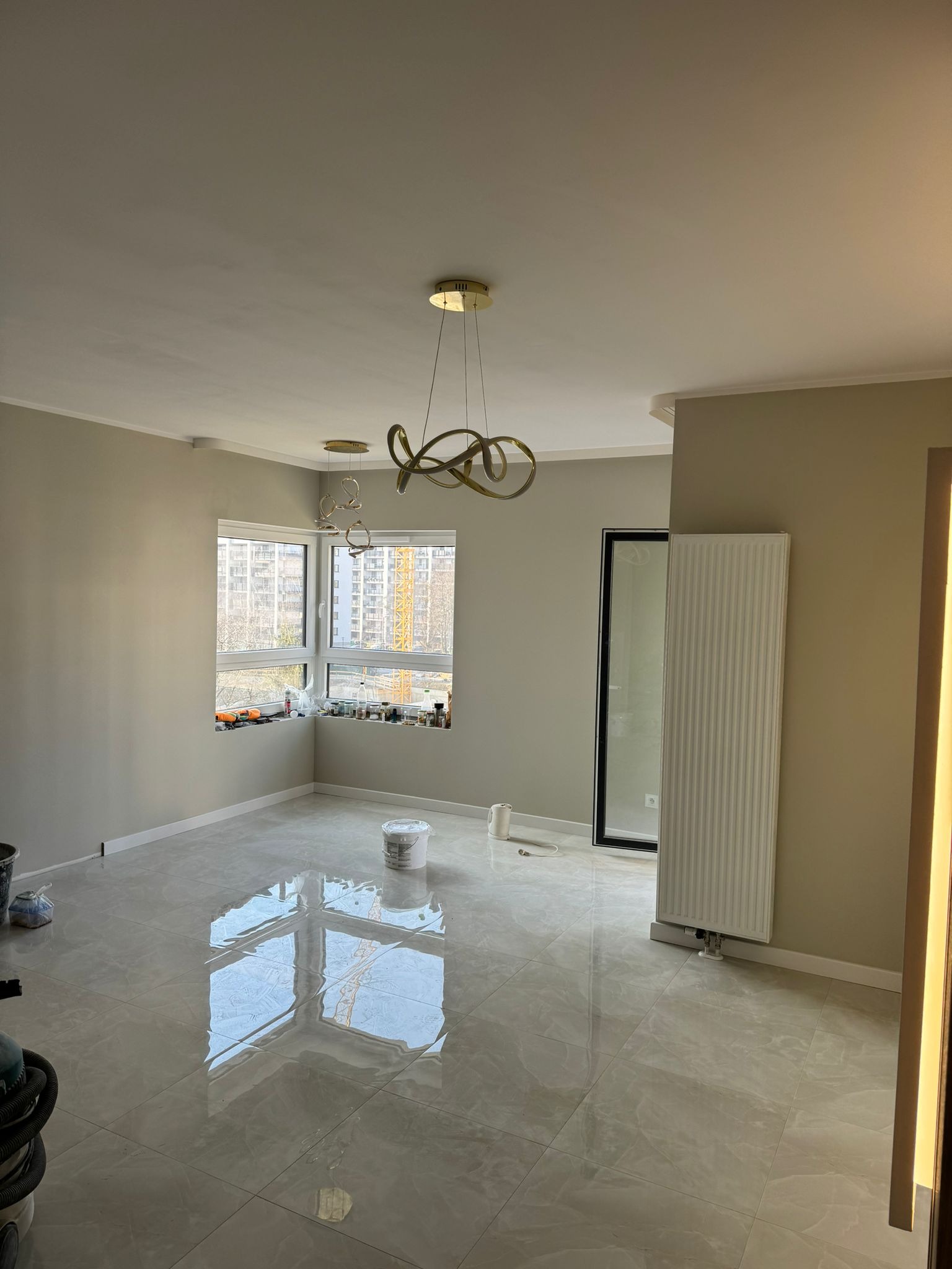 Pomalowane na szaro wnętrze pokoju z połyskującą podłogą z płytek, widoczne okno z widokiem na budowę, designerskie lampy sufitowe i biały grzejnik.