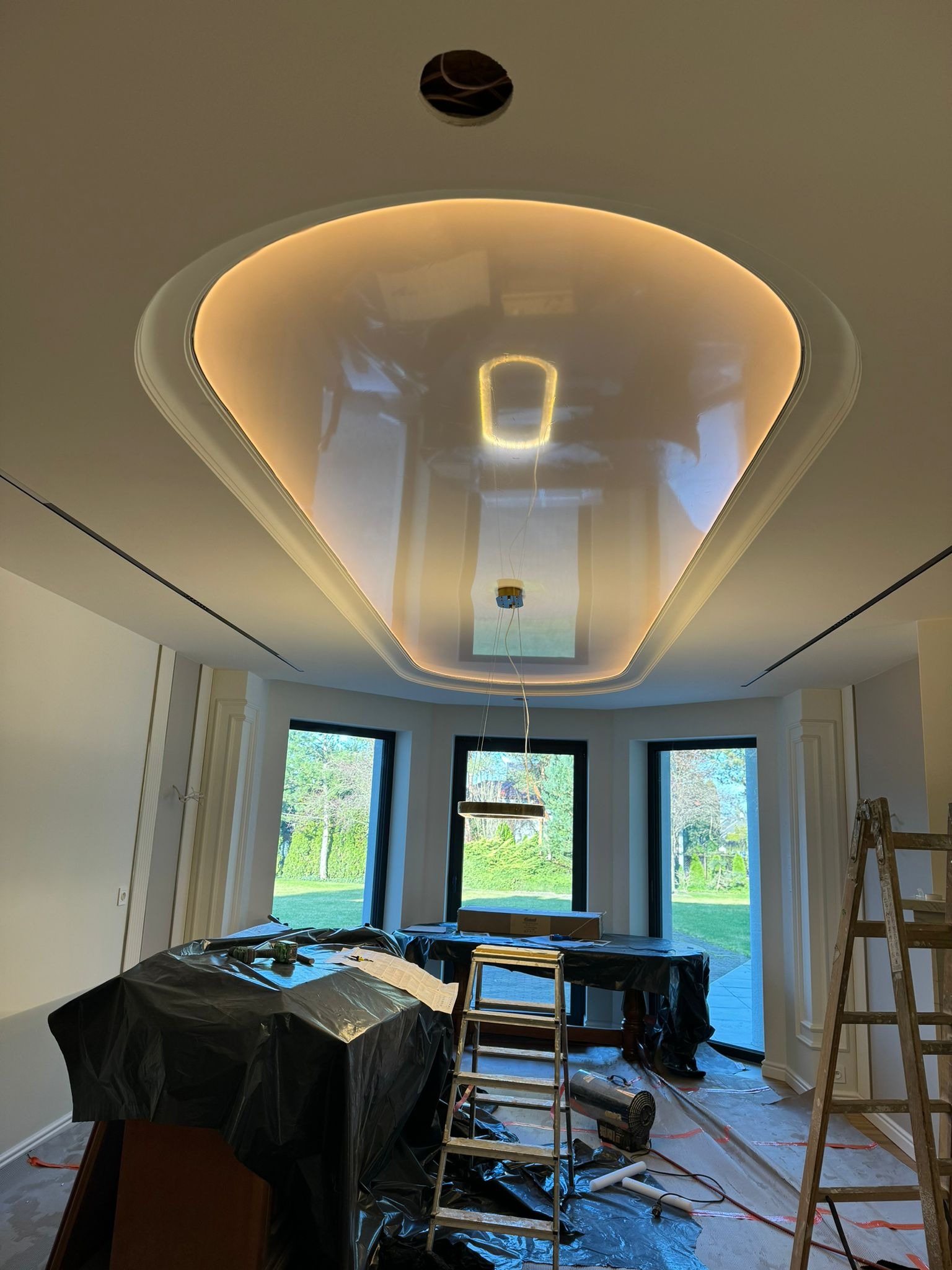 Sufit podwieszany z oświetleniem LED w kształcie elipsy podczas montażu, widoczne kable, drabina i zabezpieczone meble folią malarską.