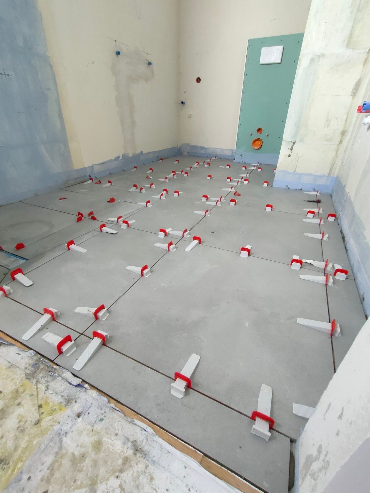Szare płytki podłogowe w trakcie układania, z użyciem poziomujących klipsów i klinów w kolorze czerwonym, widoczne ściany w trakcie przygotowania do dalszych prac.