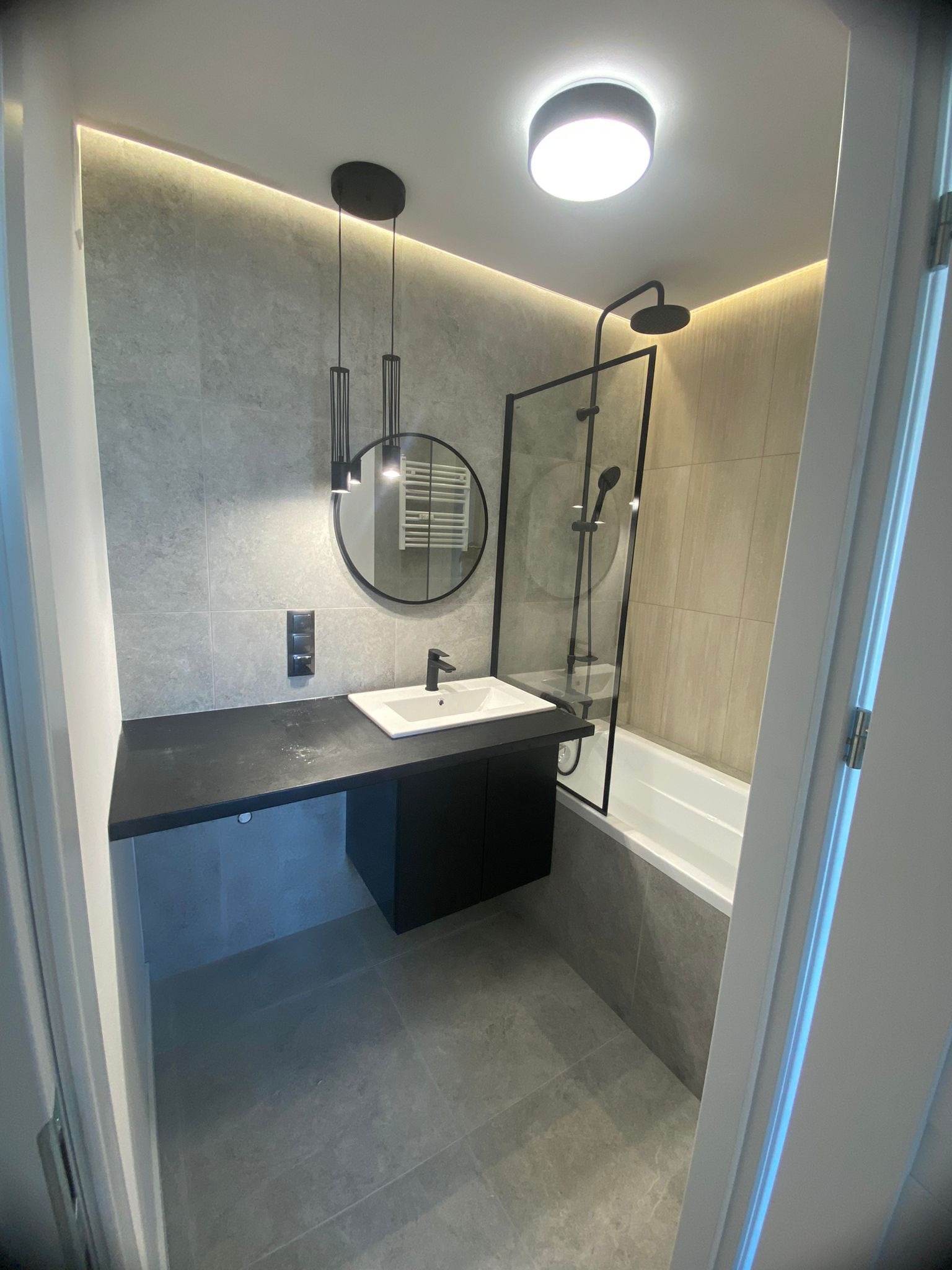 Nowoczesna łazienka z szarymi płytkami, czarnym blatem i okrągłym lustrem podwieszonym na tle podświetlenia LED, widok z wejścia przez otwarte drzwi.